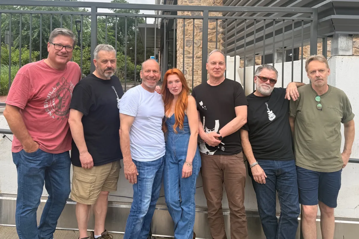 The seven band members standing together outdoors by a railing, two of them in the black guitar logo tee